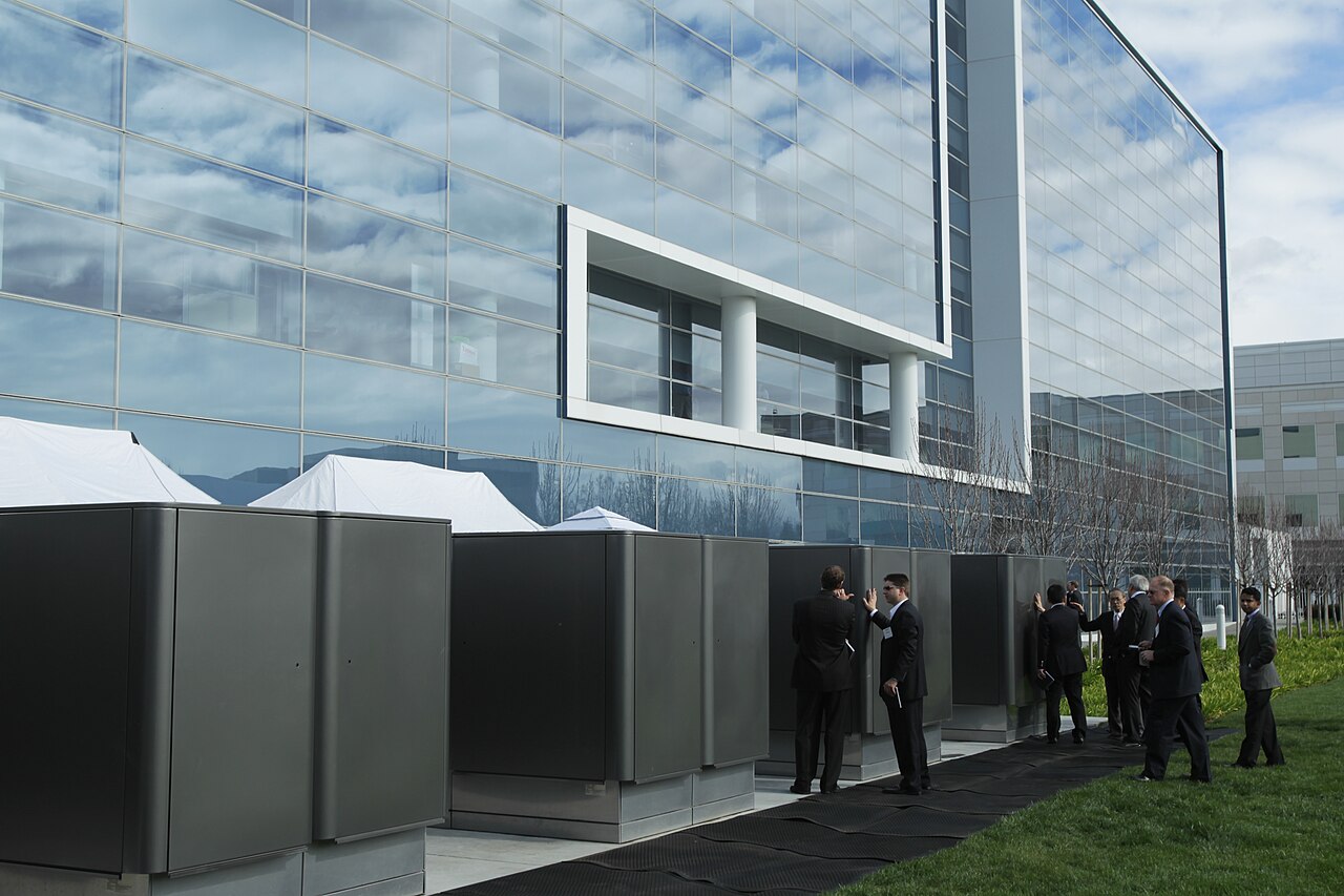 A row of grey Bloom Energy Server fuel cell cabinets beside a glass office building, with people in suits standing among them.