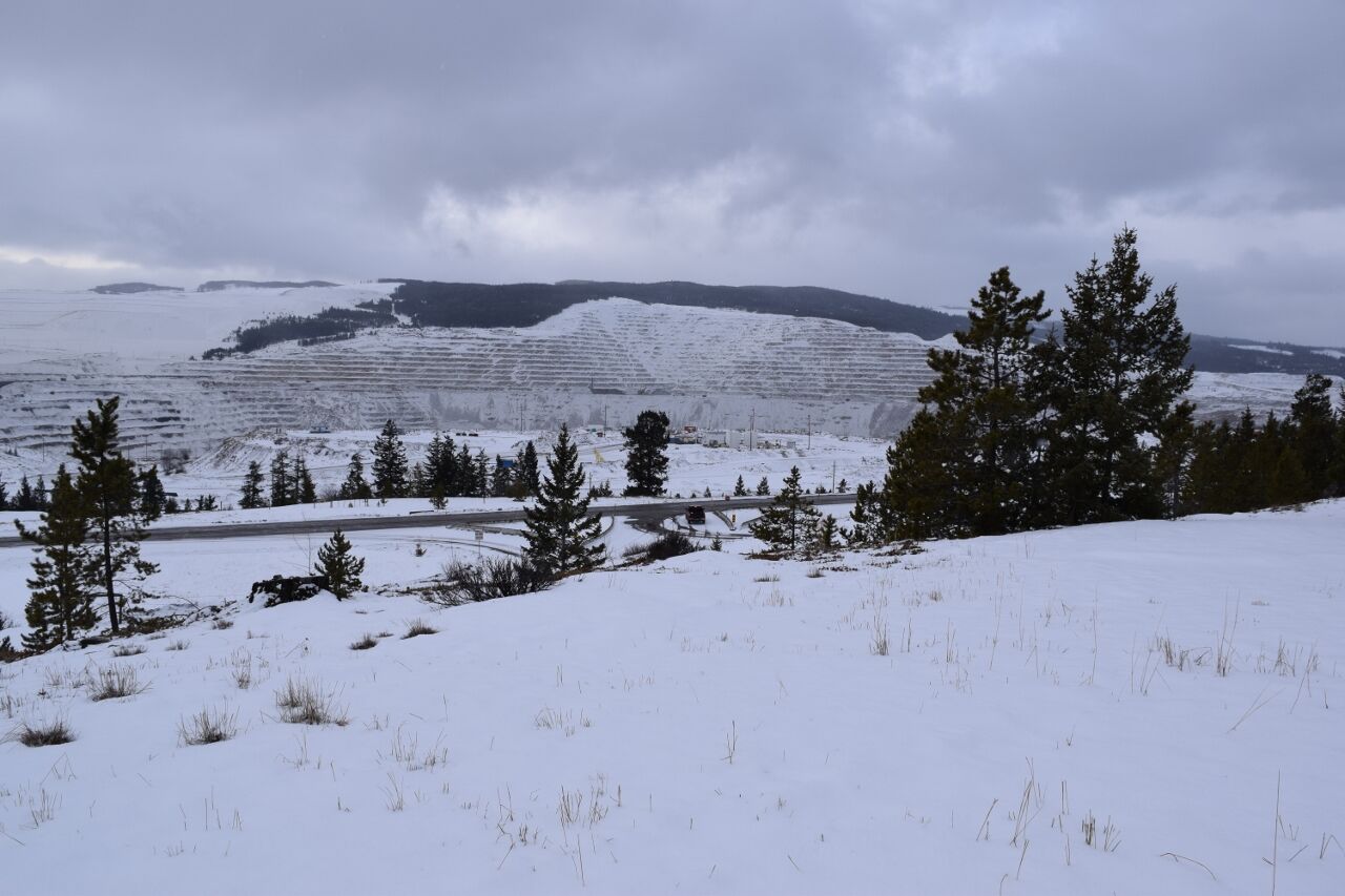 A snow-covered benched open pit wall at Highland Valley Copper under a grey sky, with pine trees and a road in the foreground.