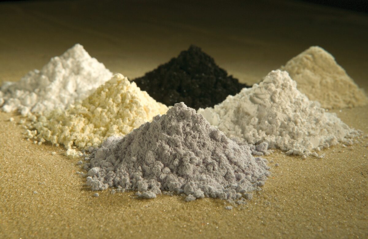 Six small piles of rare earth oxide powders on a sandy surface, in white, pale yellow, cream, grey and black.