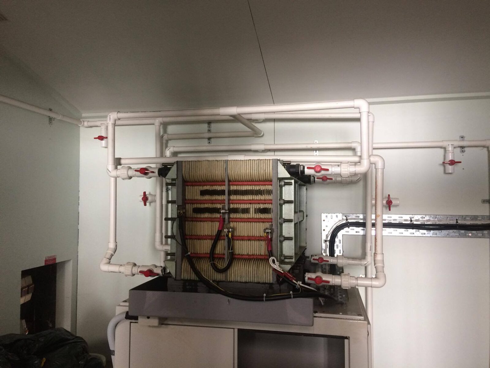 A vanadium redox flow battery stack with red cell frames, mounted on a cabinet and connected to white plastic electrolyte pipes with red valves.