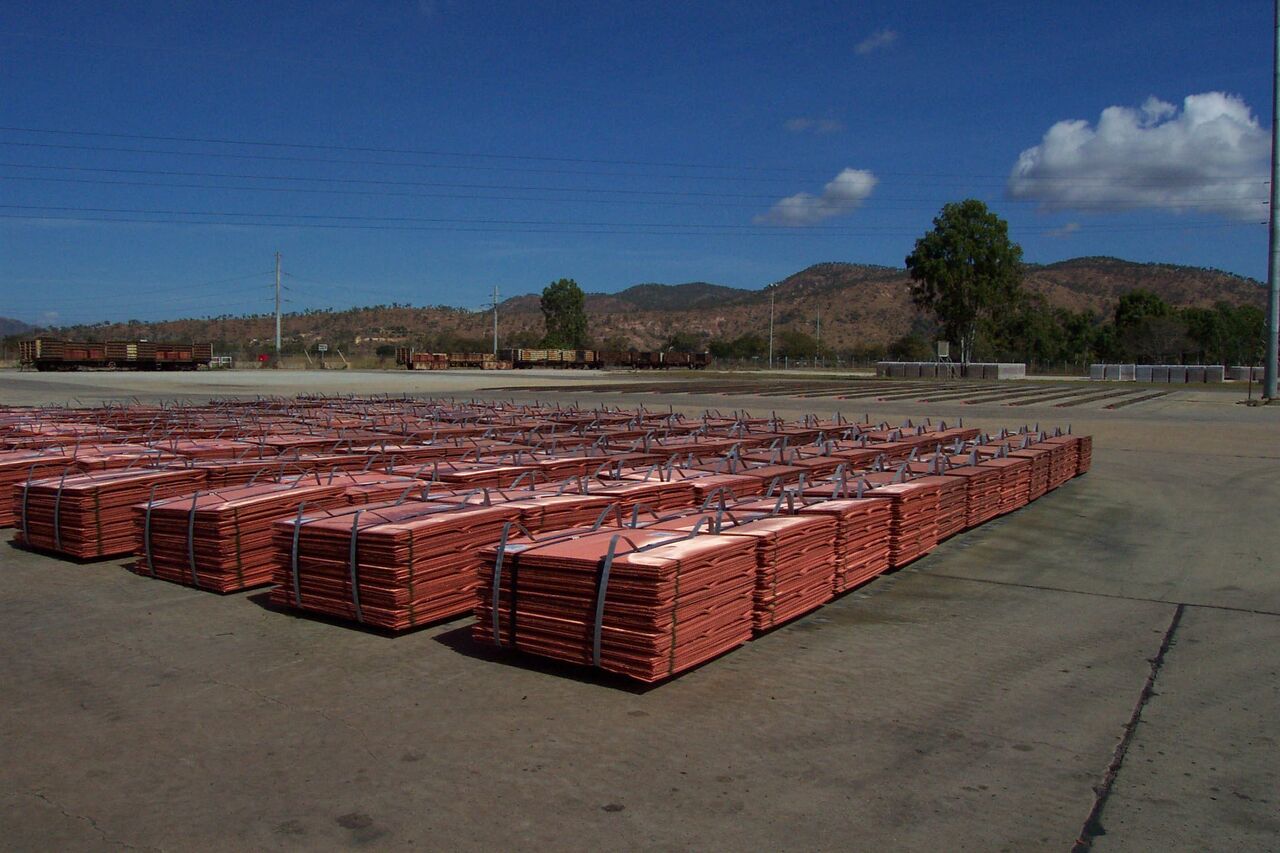 Rows of strapped bundles of copper cathode plates on a concrete yard at a refinery, with rail wagons and hills behind.