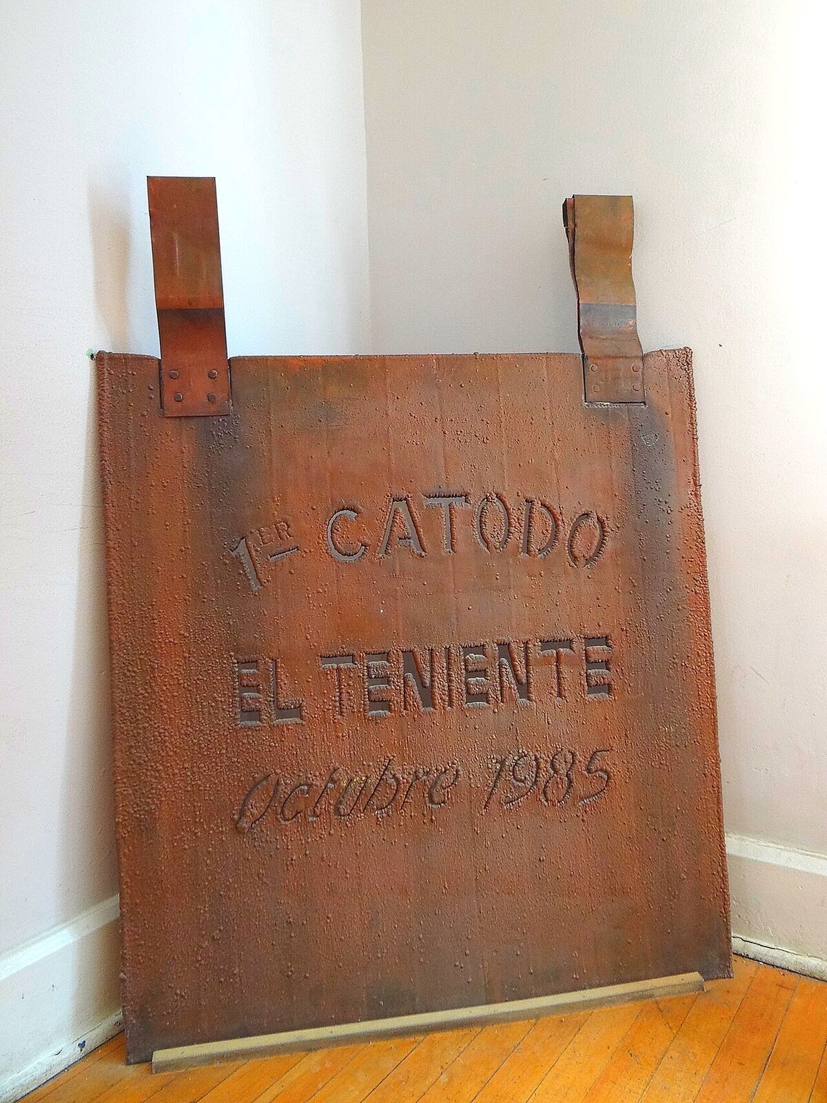 A copper cathode plate stamped "1er Catodo El Teniente Octubre 1985" leaning against a white wall in a museum corner.